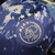 Detalhe do logotipo da camisa do Ajax II 24/25, com fundo azul marinho e verde azulado, destacando as três estrelas acima do escudo.