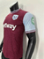Camisa do West Ham United, com detalhes em marrom e azul claro, exibindo o logotipo do time e patrocinadores.