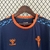 Camiseta da marca Hummel em azul escuro com detalhes em laranja e azul claro, pendurada em um cabide.