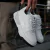 Zapatillas blancas modelo Trust One, sostenidas por una mano, con detalles en cuero y suela negra.