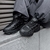 Zapatillas deportivas negras con diseño moderno, apoyadas sobre una superficie de piedra mientras una persona viste pantalones grises.