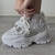 Zapatillas Aglet blancas con detalles en gris, usadas por una persona que se está atando los cordones.