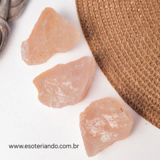 Três pedaços de quartzo rosa bruto sobre uma superfície clara ao lado de um objeto de palha.