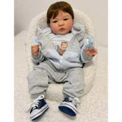 Bebê reborn menino realista sentado em poltrona branca, vestindo roupa cinza com camiseta branca e tênis azul.