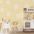 Papel de parede infantil estilo boho com flores de margarida branca em fundo amarelo claro, decorando um quarto com móveis infantis de madeira clara e brinquedos de cozinha.