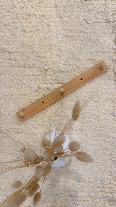 Perchero de cocina de madera con tres ganchos, sobre un fondo texturizado claro y decorado con flores secas en un jarrón blanco.