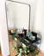 Espelho de chão retangular com moldura preta, refletindo um carrinho de bar decorado com garrafas, plantas e objetos de decoração em ambiente moderno.
