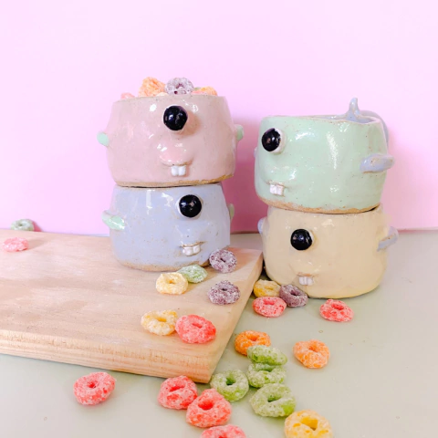 Xícaras divertidas em formato de roedor, cheias de cereais coloridos, com confeitos espalhados ao redor.