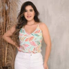 Mulher sorridente usando uma blusa colorida com estampas de folhas e calça branca, posando em frente a um fundo neutro com plantas secas.