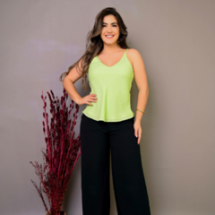 Mulher sorridente usando uma blusa verde claro e calças largas pretas, com uma planta decorativa ao fundo.