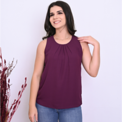 Mulher sorridente usando uma blusa roxa com plissados, posando em frente a um fundo branco.