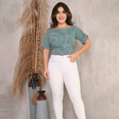Mulher posando em um fundo neutro, vestindo uma blusa listrada verde e branca e calças brancas, com decoração de plantas ao fundo.