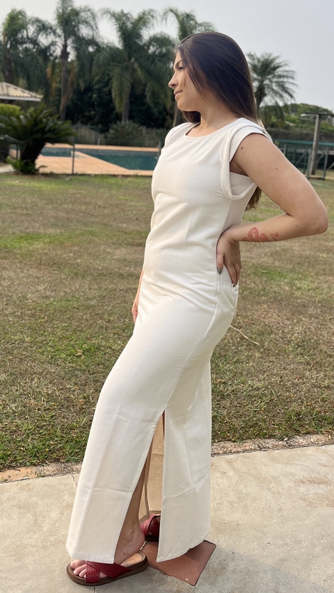 Vestido branco longo de alças largas, com fenda na lateral, posando ao ar livre em um ambiente com gramado, piscinas e árvores ao fundo.