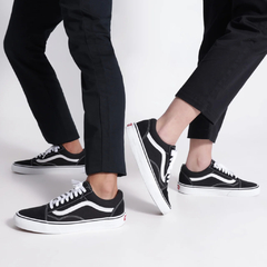 Tênis Vans Old Skool "Black" sendo usados por duas pessoas, destacando o design clássico em preto e branco.