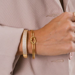 Bracelete com dois aros que se entrelaçam ao centro, formando um laço, banhado a Ouro 18k ou Ródio Branco.