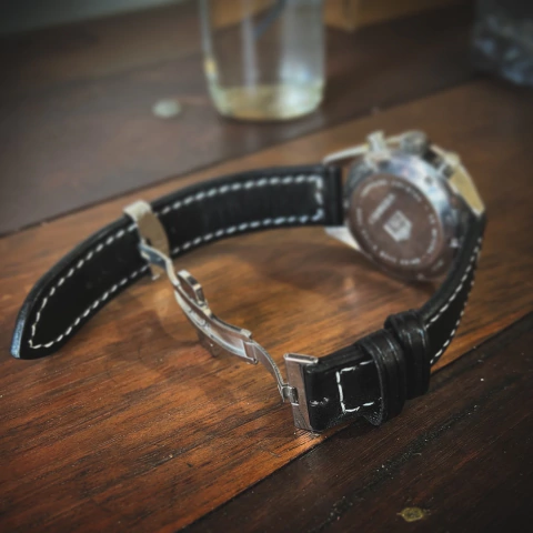 Pulseira de relógio na cor preta com fecho deployant, vista lateral sobre uma mesa de madeira.