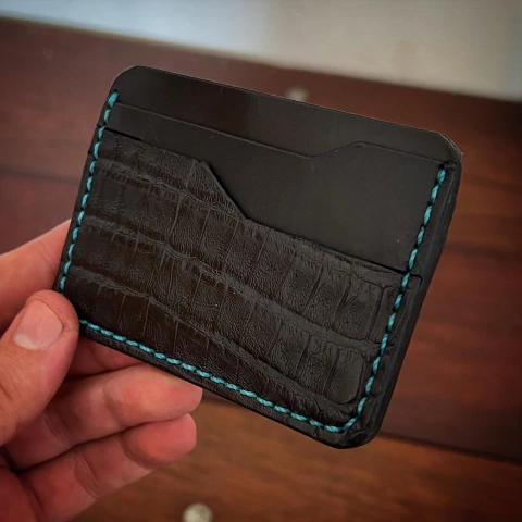 Carteira de cartão B-52 em couro de jacaré, com detalhes em costura azul, sendo segurada por uma mão.