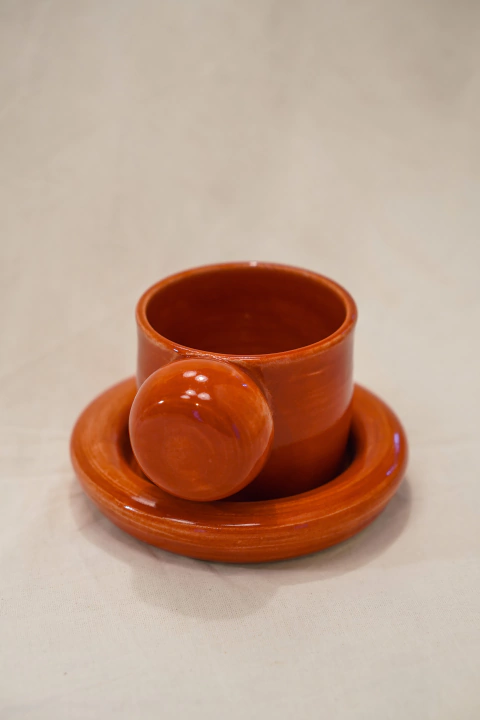 xícara de cerâmica com pires. peças são de cor vermelha. alça da xícara em formato de bola e pires com borda também arredondada. proposta de desenho para oferecer experiência de toque divertido e estética moderna.