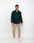 Homem sorridente usando uma jaqueta overshirt verde, com calças bege e tênis brancos, posando em um fundo branco.