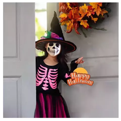 Criança vestida com fantasia infantil de bruxa para Halloween, usando chapéu pontudo preto e vestido preto com estampa de esqueleto rosa, segurando placa decorativa escrita "Happy Halloween".