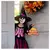 Criança vestida com fantasia infantil de bruxa para Halloween, usando chapéu pontudo preto e vestido preto com estampa de esqueleto rosa, segurando placa decorativa escrita "Happy Halloween".