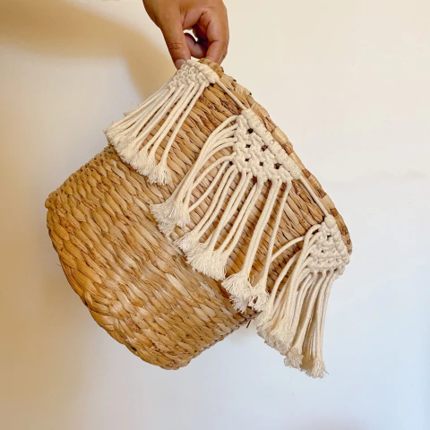 Cesto redondo de taboa com detalhe em macramê branco, sem alça, medindo 25x25 cm, sendo segurado por uma mão contra fundo branco