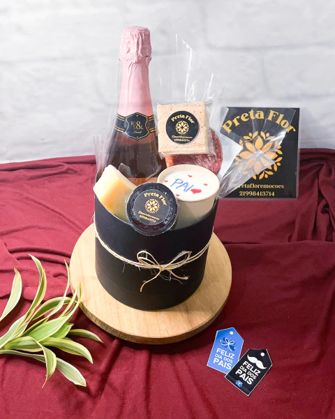 Box presente preto com garrafa de espumante, pedaços de queijo, embutidos, bolo de copo com a palavra "PAI" decorada e embalagens da marca Preta Flor