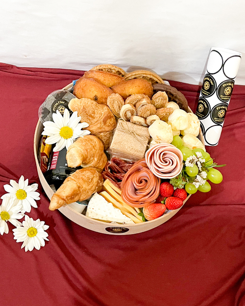 Cesta de café da manhã para 3 pessoas com croissants, pães, frios, queijos, frutas frescas e decoração floral sobre tecido vermelho
