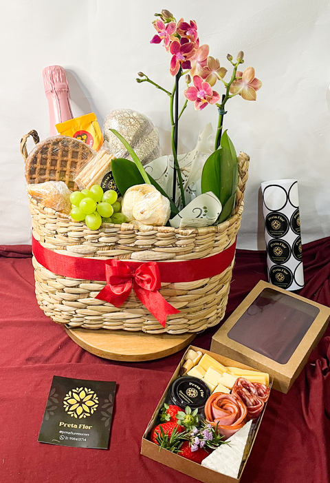Cesto gourmet decorado com flores, frutas, queijos, embutidos e uma garrafa de espumante, ideal para ocasiões românticas e celebrações.