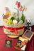 Cesto gourmet decorado com flores, frutas, queijos, embutidos e uma garrafa de espumante, ideal para ocasiões românticas e celebrações.