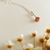 Colar longo pequeno com detalhe em cobre e corrente prateada, ao fundo com flores pequenas.