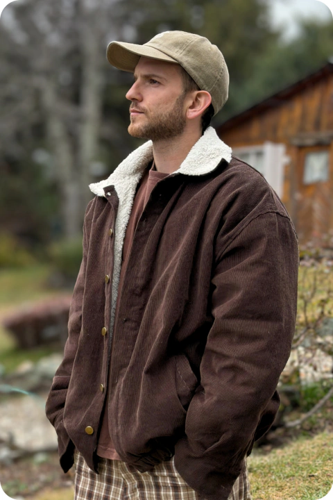 Hombre con campera de corderoy color chocolate con interior de corderito blanco, gorra beige y pantalón a cuadros en un entorno natural