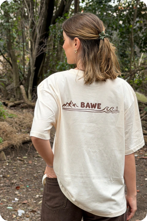 Persona de espaldas usando una remera blanca con diseño de montaña y mar en color marrón, con el texto "BAWE" en el centro