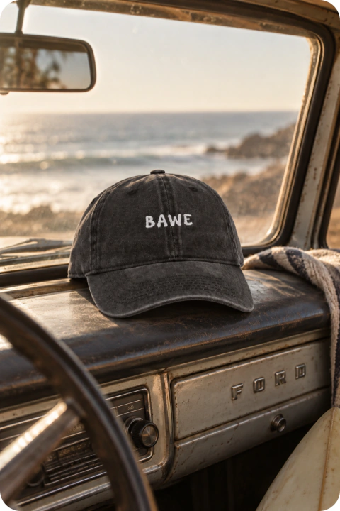 Gorra negra prelavada con logo BAWE sobre el tablero de un auto Ford antiguo con vista al mar