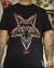 Camiseta preta com ilustração estilizada de Baphomet de quatro olhos e pentagrama invertido ao fundo, em traço vermelho e branco.