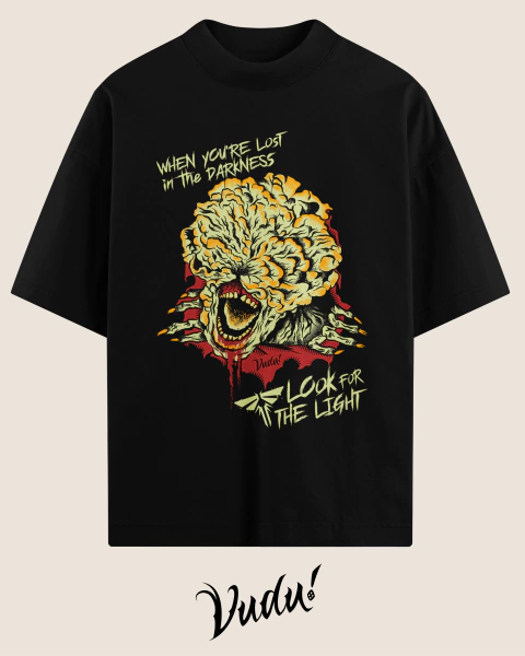 Camisa oversized preta com estampa de um Clicker do jogo The Last of Us, apresentando uma arte vibrante com a frase "When you're lost in the darkness, look for the light".