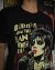 Camiseta preta com ilustração da cantora Siouxsie no microfone, com letras de músicas ao redor.