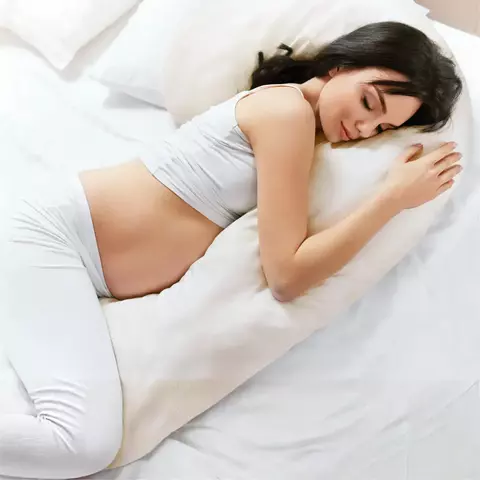Mujer embarazada descansando de lado abrazando una almohada de embarazo en forma de J color crudo