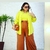 Mulher usando uma calça plus size marrom e uma blusa amarela, posando em frente a um espelho. Ela segura uma bolsa e está em um ambiente claro com uma planta ao fundo.