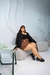Modelo usando jaqueta plus size preta, sentada em um sofá, com saia xadrez e calçados pretos.