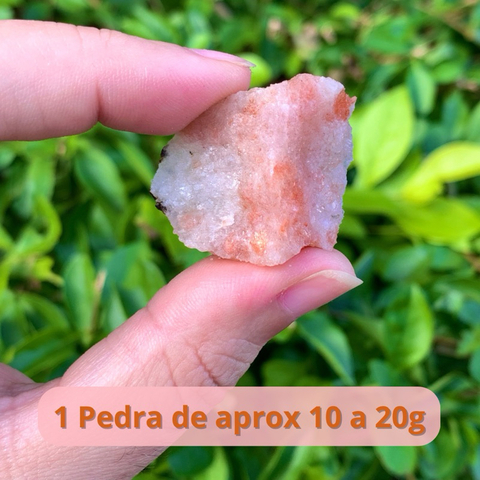 Pedra do Sol natural e bruta, com brilho, segurada entre os dedos com fundo de folhas verdes