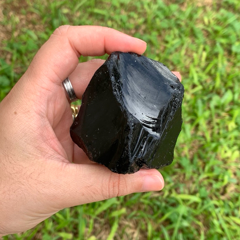 Pedra bruta de obsidiana negra natural, tamanho grande, exibida na palma da mão com fundo de folhas verdes