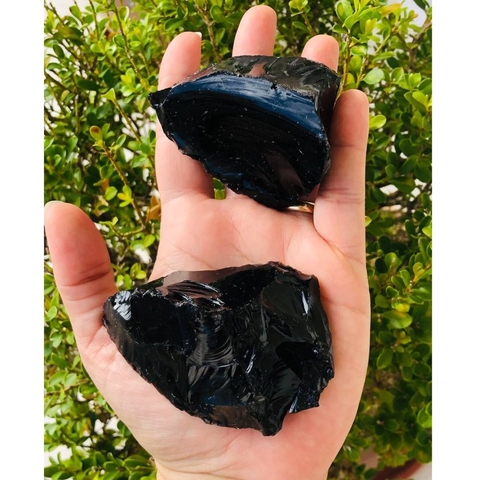 Pedra bruta de obsidiana negra natural, tamanho grande, exibida na palma da mão com fundo de folhas verdes