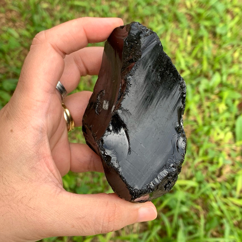Pedra bruta de obsidiana negra natural, tamanho grande, exibida na palma da mão com fundo de folhas verdes