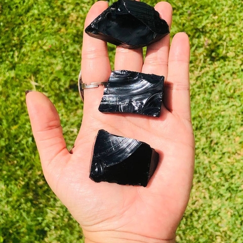 Pedra bruta de obsidiana negra natural exibida na palma da mão com fundo de folhas verdes