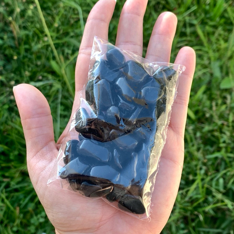 Saquinho transparente com pedras de obsidiana negra rolada, tamanho 1-2cm, segurado na mão sobre um fundo de grama verde.