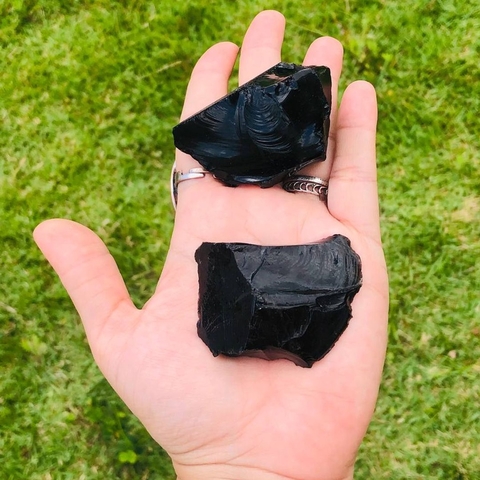 Pedra bruta de obsidiana negra natural, tamanho grande, exibida na palma da mão com fundo de folhas verdes