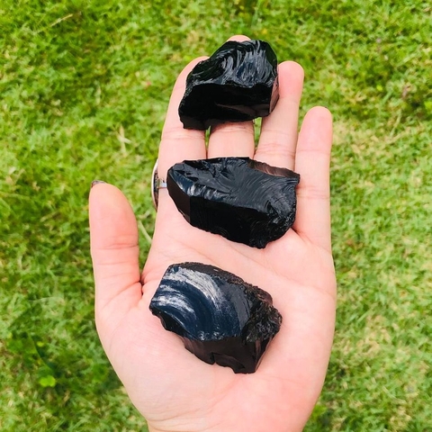 Pedra bruta de obsidiana negra natural,  exibida na palma da mão com fundo de folhas verdes