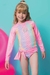 Menina usando um conjunto de biquíni e blusa em malha cirrê, com proteção UV 50+, em cores pastel, sorrindo para a câmera.
