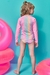 Menina de costas vestindo um conjunto de biquíni e blusa em malha cirrê, com proteção UV 50+, em um fundo colorido com boias infláveis ao redor.
